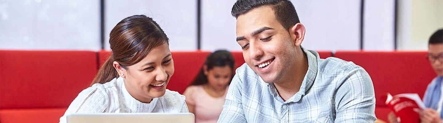 Two people are sitting together, smiling and looking at a tablet. The woman on the left has dark hair tied back, and the man on the right has short dark hair. In the background, another person is reading a book. They are seated indoors.