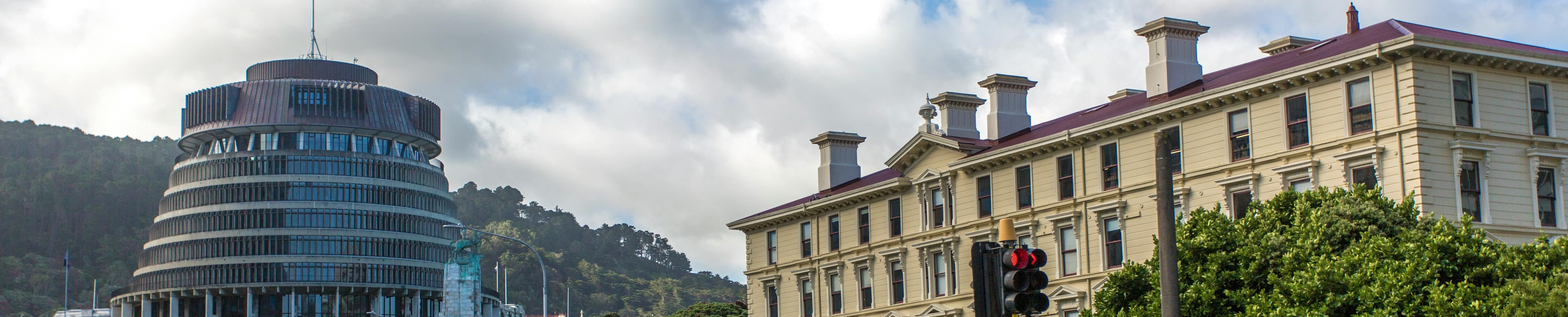 Panoramic view of a modern circular building and a historic stone building under a partly cloudy sky, surrounded by greenery and traffic lights.